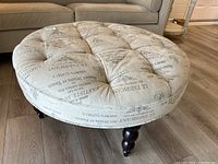 Round beige tufted ottoman with black printed text on fabric upholstery, showing the top surface and tufting detail.