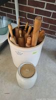 Large beige stoneware crock holding multiple vintage wooden rolling pins against a brick wall background.