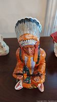 Front view of Royal Doulton figurine depicting a Native American chief in an orange outfit and feathered headdress, holding a rifle.