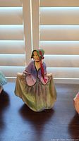 Photo of the Royal Doulton Sweet Anne porcelain figurine seated, wearing a bonnet and a purple jacket with a green skirt.