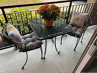 Front view of the wrought iron bistro set showing the table, two chairs with cushions, and the potted plant on the table.