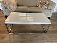 Front view of coffee table placed in front of a beige couch, showing white glossy laminate top and metal frame base.