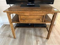 Front view of the antique wood table with a TV on top. Shows the single center drawer and two open shelves below.