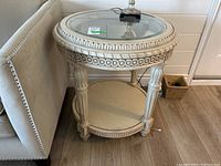 Round wood veneer side table with antique finish, inset glass top, carved details on the rim and legs, and a lower circular shelf. Positioned next to a beige upholstered couch on a wood floor.