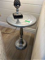 Metal side table showing top and pedestal base from a frontal angle, placed on a wooden floor next to a wall and a beige piece of furniture.