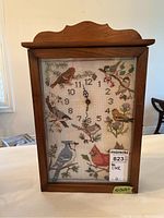 Front view of wooden wall clock showing needlework bird design and clock hands at 11:03.