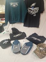 Two XL Eagles hooded sweatshirts displayed, one black and one green, plus a black NFL Champions T-shirt, two Eagles logo hats, and a pair of slippers with Eagles logo.