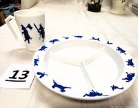 Photo showing the milk glass mug with blue silhouettes of the Three Little Pigs and the Wolf alongside the divided child’s dish with blue animal silhouettes around the rim on a white fabric background.
