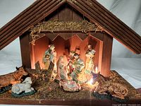 Ceramic nativity scene figurines arranged inside wooden creche illuminated by small lights beneath.