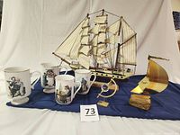 Wide view of tall ship model, crystal caliper, and four Norman Rockwell mugs on blue cloth