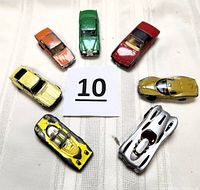 Top view of seven vintage die-cast cars arranged in a semi-circle, showing various colors and paint wear on a white knit fabric background.