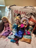 Four Disney princess plush dolls sitting side by side on white surface, with a Mulan shirt draped behind them, showing the dolls' detailed outfits and colors clearly.