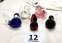 Five assorted hand-blown glass birds grouped on a table with white cloth, showing different colors and clear bases.