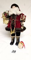 Full front view of 18-inch standing Santa figurine with ceramic face, hands, black and gold boots, velvet coat with brown fuzzy trim, gold bag and wrapped gifts, brass instrument in hand, and separate Santa head ornament placed beside it.