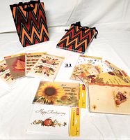 Thanksgiving greeting cards arranged in fanned sets showing various autumnal designs including turkeys and sunflowers; two gift bags with chevron zig-zag pattern in orange, black, and red with black ribbon handles