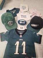 Photo of Philadelphia Eagles toddler apparel and accessories laid out including three onesies in different colors, a burp cloth, two toddler hats, a 12-month jersey with number 11, and a soft football.