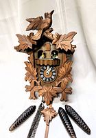 Front view of vintage wooden cuckoo clock showing carved bird, leaves, dancing figurines, clock face with Roman numerals, and pinecone weights.