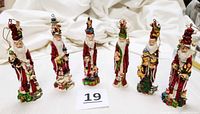Front view of six vintage pencil Santa figurines arranged in a row, showing detailed holiday accents including elves, snowmen, toy soldiers, angels, toys, and teddies. Each approximately 6 inches tall with bright red clothing and decorative bases.