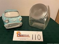Both items in the lot: Applause Wheelers ‘57 Chevy cookie jar and large clear glass counter display jar with hinged metal lid, sitting on a green tablecloth.