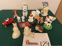 Wide shot showing entire lot on green cloth background with white wall behind. Items include various animal figurines, Disney Mickey Mouse in red car, salt and pepper shakers, and novelty banks.