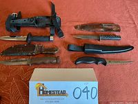 Top-down view of four knives arranged with their respective sheaths on a red patterned fabric surface with lot card.
