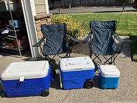 Two blue folding camp chairs with armrests and storage bags, positioned next to three coolers — large blue Coleman rolling cooler, medium blue Igloo rolling cooler, and small blue and white Ice Cube cooler, outside on a driveway.