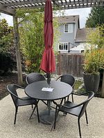 Photo of the round folding table, four matching chairs, and red closed umbrella on a patio surface under pergola.