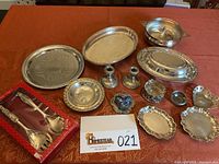 Overview of all silverplate items laid out on orange tablecloth, including trays, candlesticks, salad set in box, various decorative dishes and covered serving piece