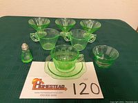 Nine-piece set of green Vaseline glassware with etched parrot and palm tree designs displayed on a black cloth with lot number card.