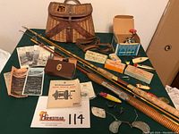 Photo showing entire vintage fishing collection including fishing poles, reels, creel, assorted lures, and pamphlets laid out on green table.