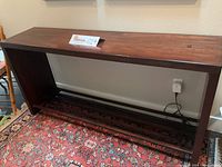Wooden sofa table showing top and sides with dark finish, placed against a wall with an outlet and carpeted floor.