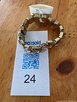 Faux smokey quartz costume bracelet by Joe shown resting on a MaxSold tag. The elasticized band and polished gold-tone segments are visible with a large central faux stone.