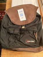 Front view of black canvas Eastpak backpack with brown leather flap and bottom, showing Eastpak logo patch on front pocket.