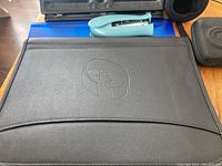 Photo showing a blue three hole punch, a light blue metal stapler, and a large black portfolio folder with embossed logo.
