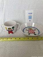 Kids cup and bowl set placed side by side on white textured surface with measuring tape showing dimensions.