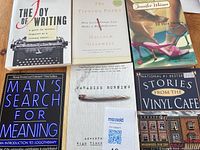 Six paperback books arranged on a wooden surface. Titles visible: The Joy of Writing, The Tipping Point, In Her Shoes, Man's Search for Meaning, Paradise Burning, Stories from the Vinyl Cafe. Covers show good condition with minor signs of use.