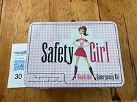 Top view of the Safety Girl Roadside Emergency Kit tin case with branding and illustration.