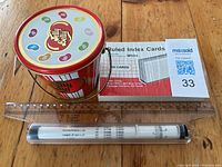 Photo showing Jelly Belly branded metal tin, ruled white index cards, clear plastic ruler, and cylindrical slide rule case on wooden surface.