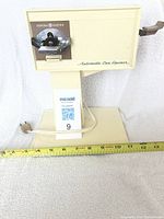 Front view of the vintage beige General Electric automatic can opener on its stand with a visible measuring tape at base, showing size.