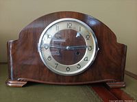 Front view of the wooden mantle clock showing rounded top, polished wood grain, round dial with Arabic numerals, and glass cover.