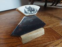 Front view of the ulu showing the stone handle etched with a moose and forest scene, and the broad stainless steel blade resting on a wooden stand.