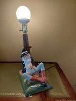 Side angle of vintage 1950s chalkware streetlamp with seated old man figure holding a shoe, lamp lit.