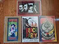 Four vintage concert posters laid out on wood floor, each in protective vinyl sleeves. Whitish border edges visible around posters.