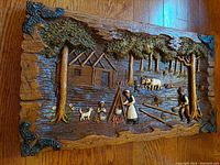 Photo of vintage carved wood diorama with pioneer figures, cabin, trees, campfire, dog, and cow, with decorative metal corner brackets, on wood floor.