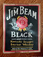 Front view of wooden plank showing Jim Beam Black hand painted sign featuring logo and text with visible wood grain.