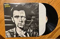 Front cover of Peter Gabriel vinyl record album showing black and white close-up portrait with half the face distorted, record partially visible inside white protective sleeve.