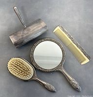 Photo showing all four items: silver plated crumb sweeper with hinged top and handle, handheld mirror, hairbrush with cream bristles, and metal decorative comb arranged on flat surface.