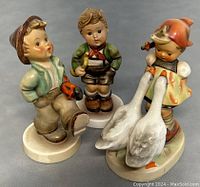 Front view of three hand-painted Hummel figurines featuring children in traditional Bavarian clothing with detailed colors and two white geese.