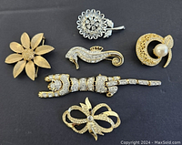 Top view of six vintage brooches arranged on black background showing various gold-tone and silver-tone designs with rhinestones and faux pearl accents.