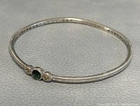 Full side-angle view of silver bangle showing ribbed texture and green stone and two clear stone accents on front middle.